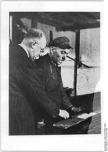 "Die Verbesserungsvorschläge der Werktätigen, in Aktivistenplänen zusammengefaßt, sind für die vorfristige Erfüllung des 5-Jahrplanes von ausschlaggebender Bedeutung, da sie Rohstoffe, Zeit und Geld einsparen helfen. Der Betriebsleiter Gansauge von den Elite-Diamant-Fahrradwerken in Chemnitz entwickelte eine Meßlatte, um den bisher beträchlichen Abfall an Rohrenden zu beseitigen. Die Rohre für Fahrradrahmen die in ihrer Länge sehr verschieden sind, werden mit Hilfe dieser Latte aufs rationellste ausgenutzt. Hatten die Abfallstücke bisher eine durchschnittliche Länge von 80 cm, so bleiben jetzt nur noch Reste von höchstens 30 cm übrig. Die Jahrseinsparung beträgt ca. 4.000 DM. Betriebsleiter Gansauge (links) erklärt einem Arbeiter im Rohrlager das Abstechen der Rohre mit Hilfe der Meßlatte." (Februar 1951)