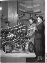 "Im Pavillon vor dem Hause des Ministeriums für Finanzen läuft noch bis Sonntag die Ausstellung: "Fünf Jahre Aufbau der Friedenswirtschaft im demokratischen Sektor von Berlin". Die Ausstellung hat beim Publikum bisher grossen Anklang gefunden. Frau Fordan und Frau Margarete Müller (vlnr) betrachten die von der Firma Woitschach, Bln.-Karow gefertigten "Teddy-Sporträder"." (Oktober 1954)