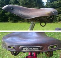 Sportsattel von ?? Zeitraum: 195x bis 1957 Verwendung: Zubehörteil für Simson-Fahrräder Lederdecke: braun lackiert Gestell: verchromt Bemerkungen: lange Ausführung, Gestell mit zusätzlichen Federschleifen, Simson-Plakette aus Kunststoff an der Rückseite